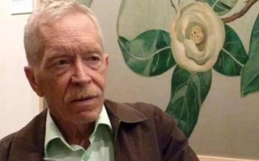 An older Caucasian man with a white moustached tinged with ginger wears a green shirt and brown jacket. He is looking off to the right and cradling a paper coffee cup.